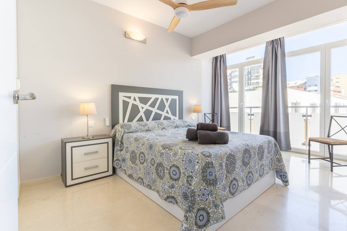Une chambre à coucher avec un lit, deux lampes de chevet, un ventilateur de plafond et une vue sur les bâtiments à travers de grandes fenêtres.