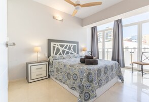 A bedroom with a bed, two bedside lamps, a ceiling fan, and a view of buildings through large windows.