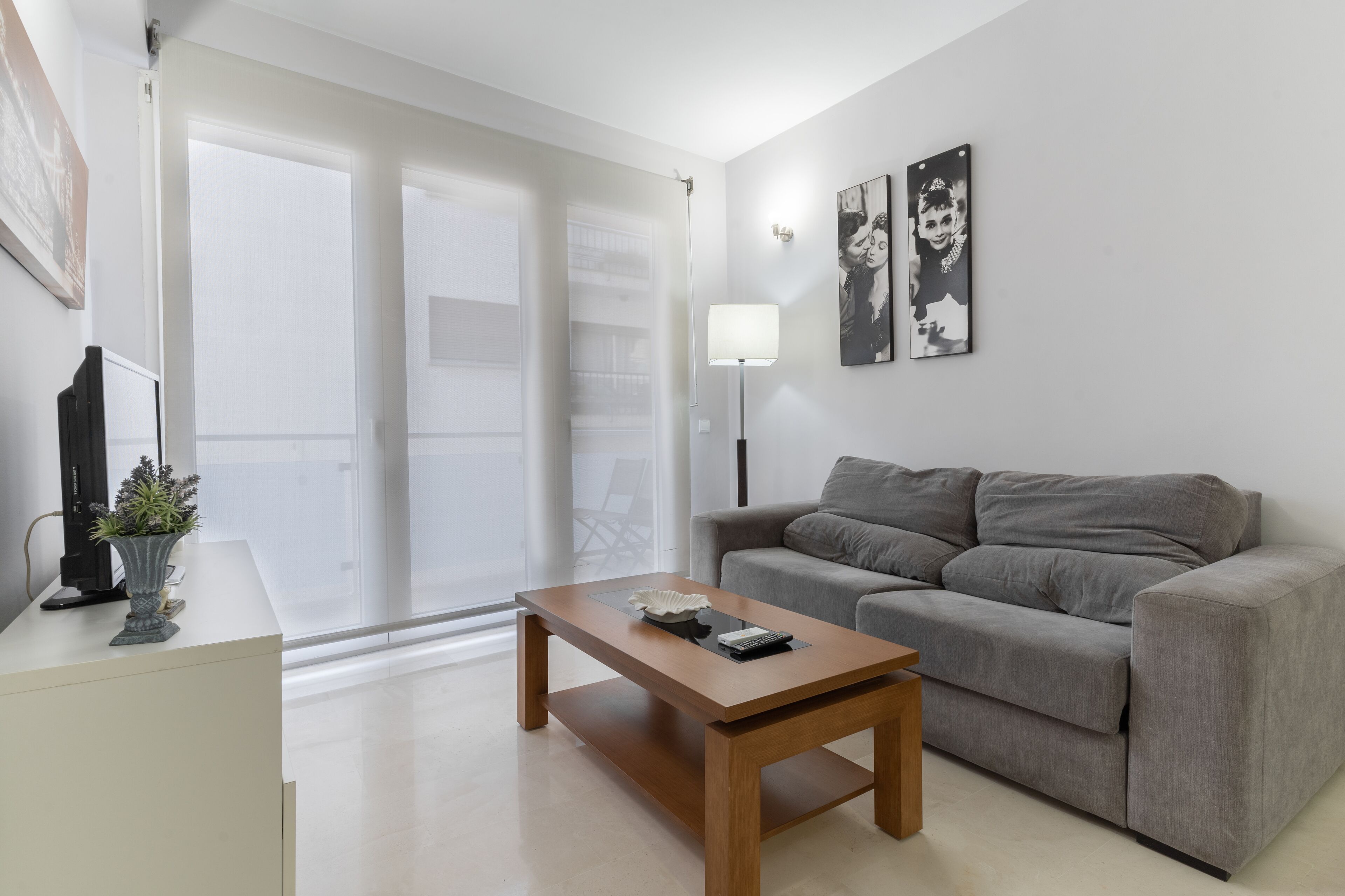 A modern living room with a grey sofa, a wooden coffee table, and a floor lamp. There is a large window with blinds, a potted plant, and a flat-screen TV on a stand.