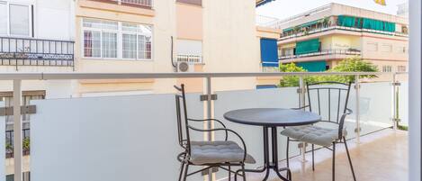 A balcony with two metal chairs and a small round table, overlooking a multi-story building with balconies and a yellow awning.