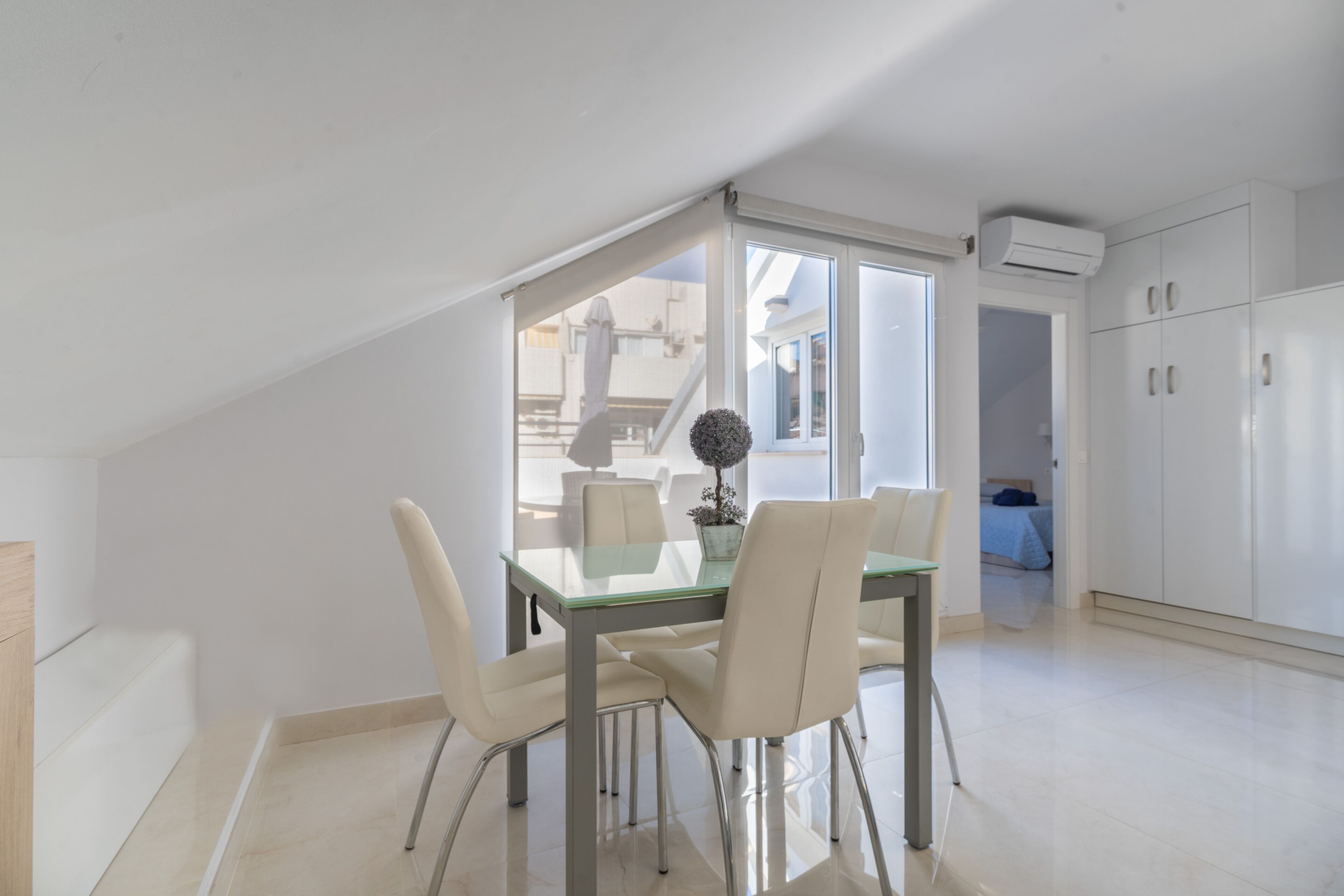 A modern, well-lit dining area with a glass table and white chairs, leading to a bedroom and a kitchen area.