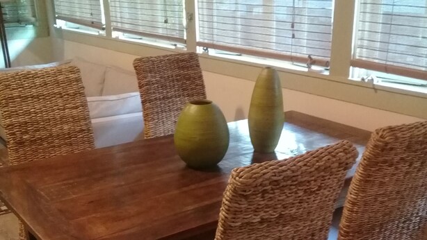 Une table à manger entourée de chaises en osier, deux vases et une fenêtre avec des stores.