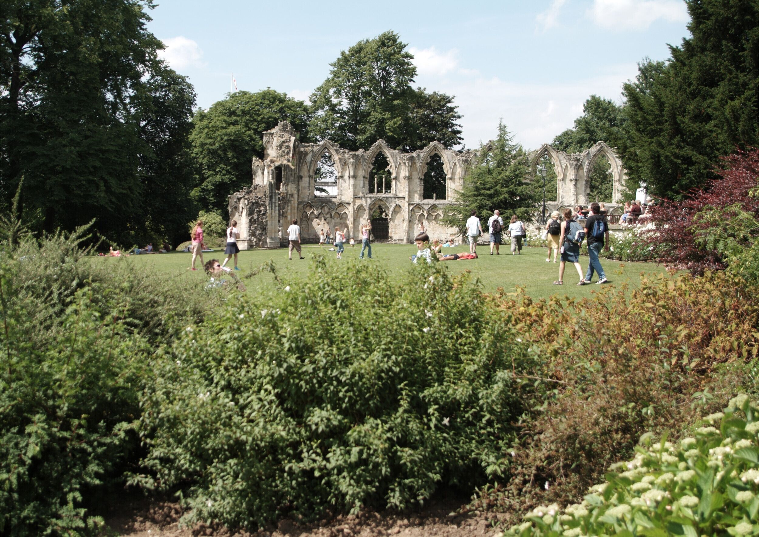 An ancient stone structure with Gothic arches surrounded by lush greenery and visitors.