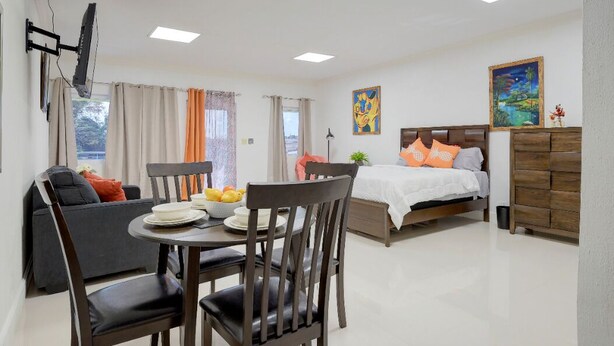 A modern hotel room with a dining area, a bed with orange pillows, and artwork on the walls. - The Ideal Loft (Nassau)