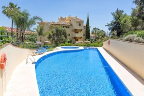 A swimming pool with lounge chairs and a lifebuoy, surrounded by palm trees and a building with a tiled roof.