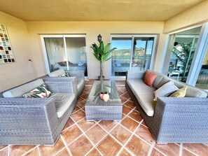 A spacious living area with wicker furniture, a glass coffee table, and a large sliding glass door leading to a balcony with a view.
