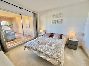A bedroom with a large bed, two bedside tables, a sliding glass door leading to a balcony, and a wall with three framed artworks.