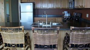 Una cocina con armarios de madera, refrigerador de acero inoxidable, pileta doble y taburetes de mimbre.