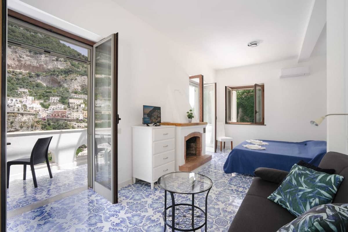 A bedroom with a balcony, a bed, a dresser, a small table, and a fireplace.