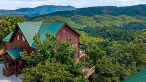 A log cabin with a green roof, surrounded by lush greenery and mountains in the background. - Beaver's Treehouse - Private Pool and Sauna! (Pigeon Forge)