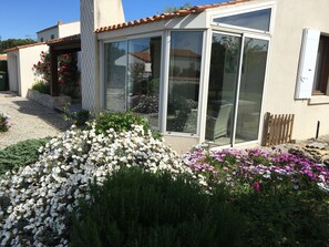 A house with a patio area, a garden with white and purple flowers, and a sliding glass door. - Quiet house in a private dead end 150m from the beach. (Saint-Denis-d'Oléron)