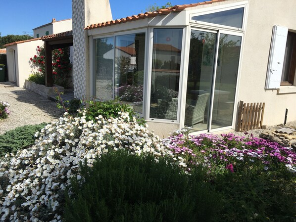 A house with a patio area, a garden with white and purple flowers, and a sliding glass door. - Quiet house in a private dead end 150m from the beach. (Saint-Denis-d'Oléron)