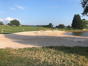 Ett sandstrandområde med ett volleybollnät, en liten damm och en gräsklädd förgrund.