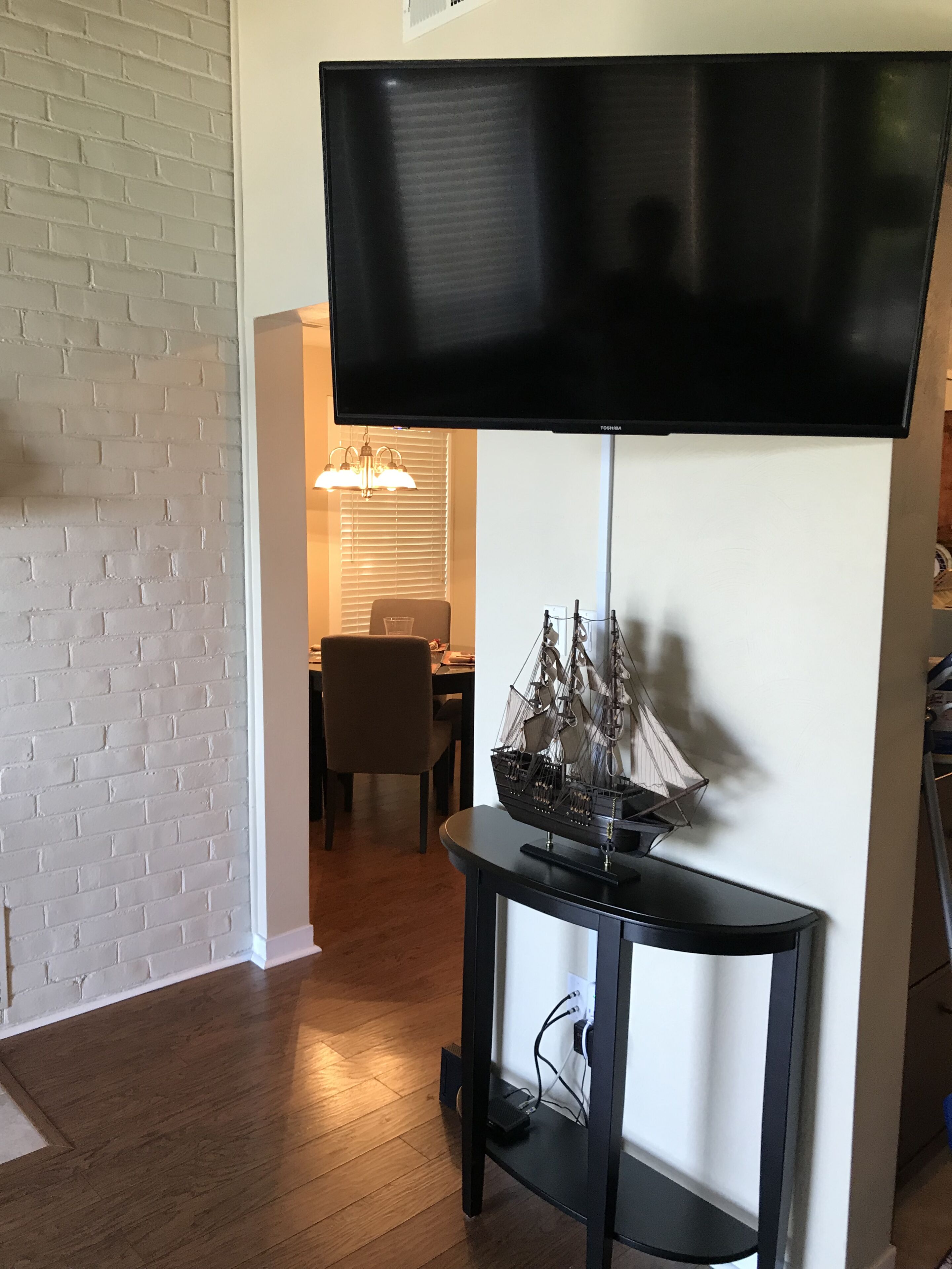 A living room with a flat-screen TV mounted on a black stand, a ship model on a side table, a brick-patterned wall, and a dining area in the background.