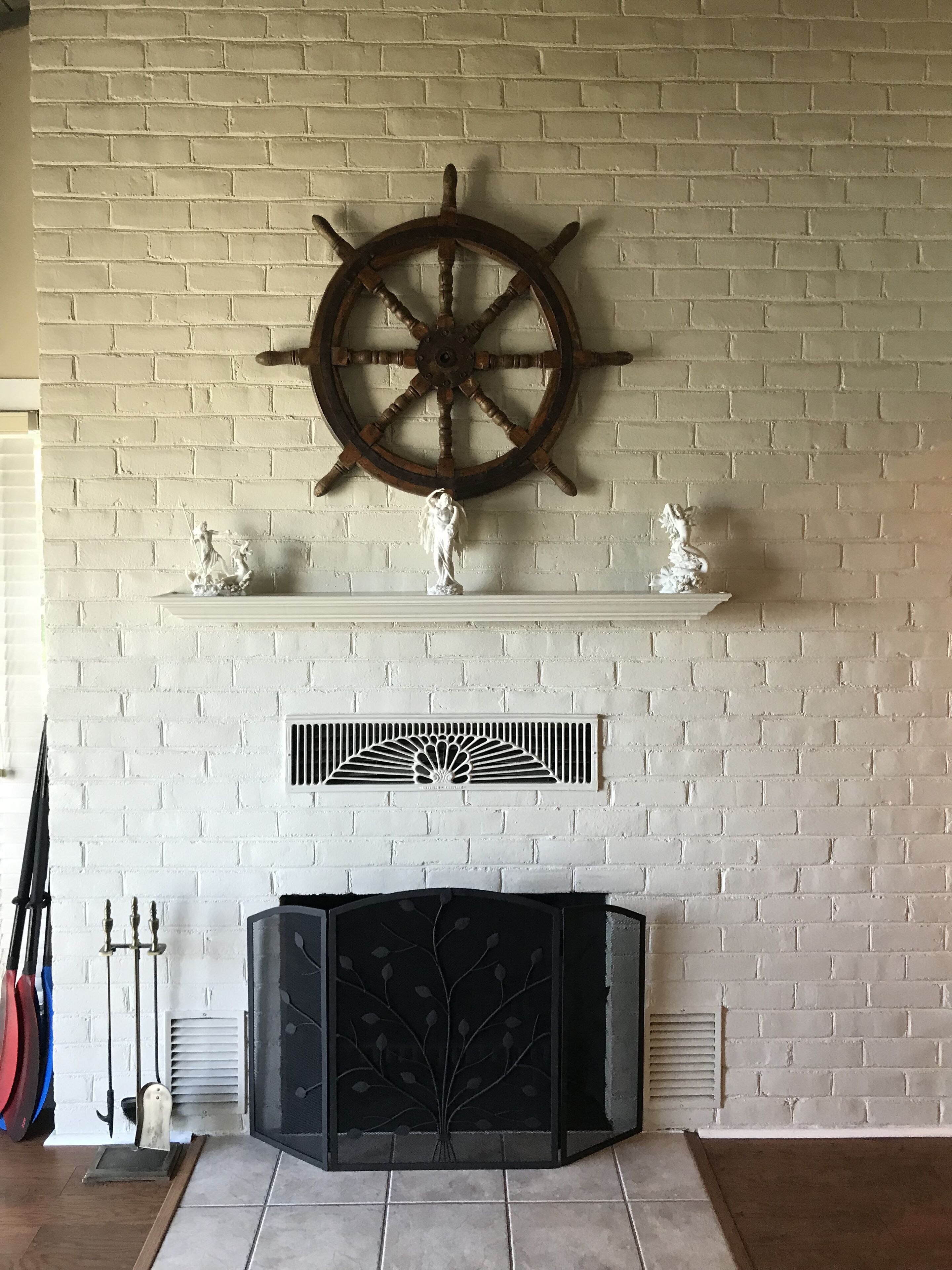A fireplace with a decorative mantle, a ship's wheel above it, and a white brick wall.
