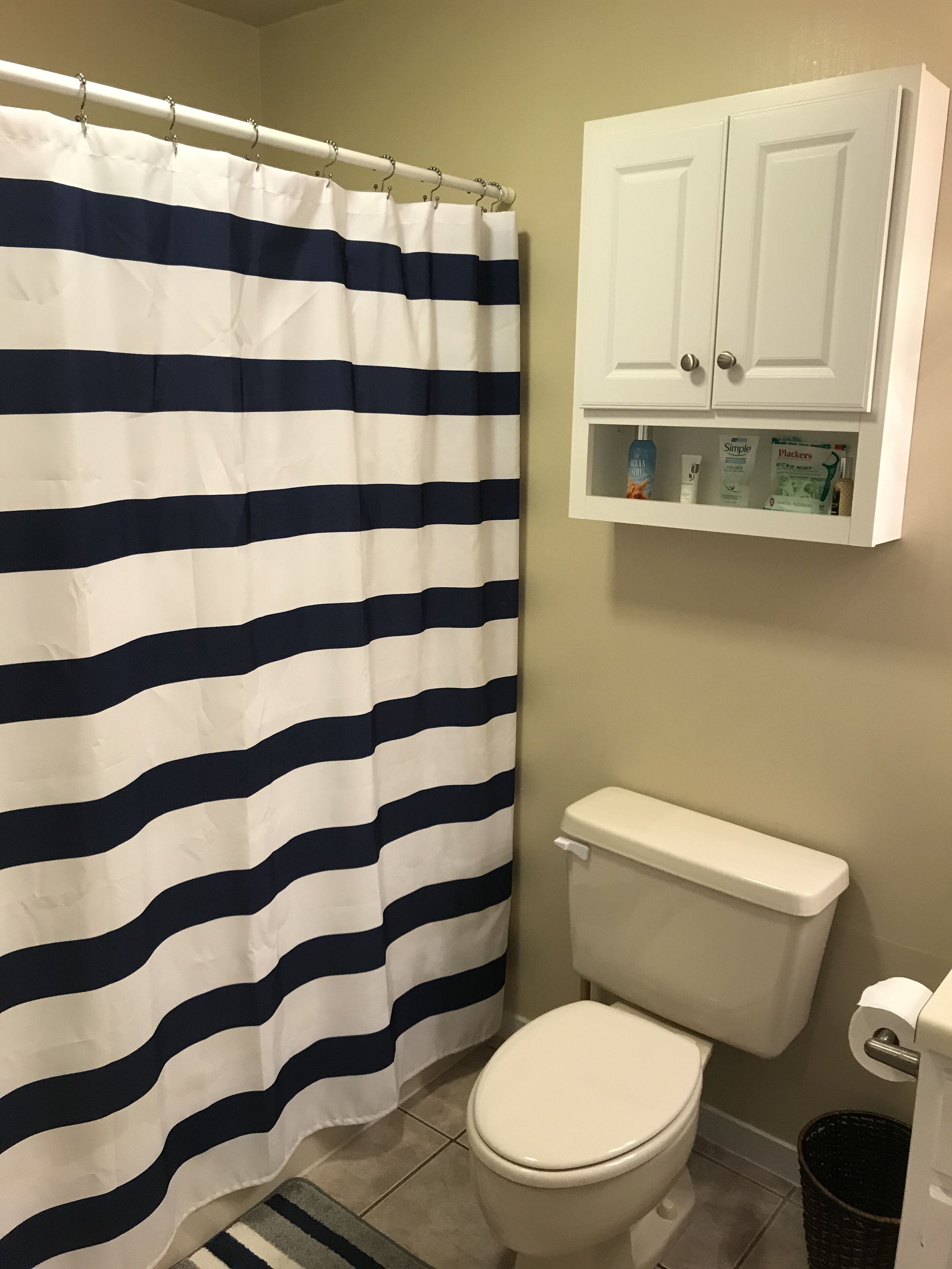 A bathroom with a striped shower curtain, a toilet, a cabinet with toiletries, and a trash bin.