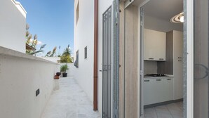 Un estrecho pasillo exterior con una pared blanca, plantas en macetas y una reja metálica que conduce a una zona de cocina con armarios blancos y suelo de baldosas.