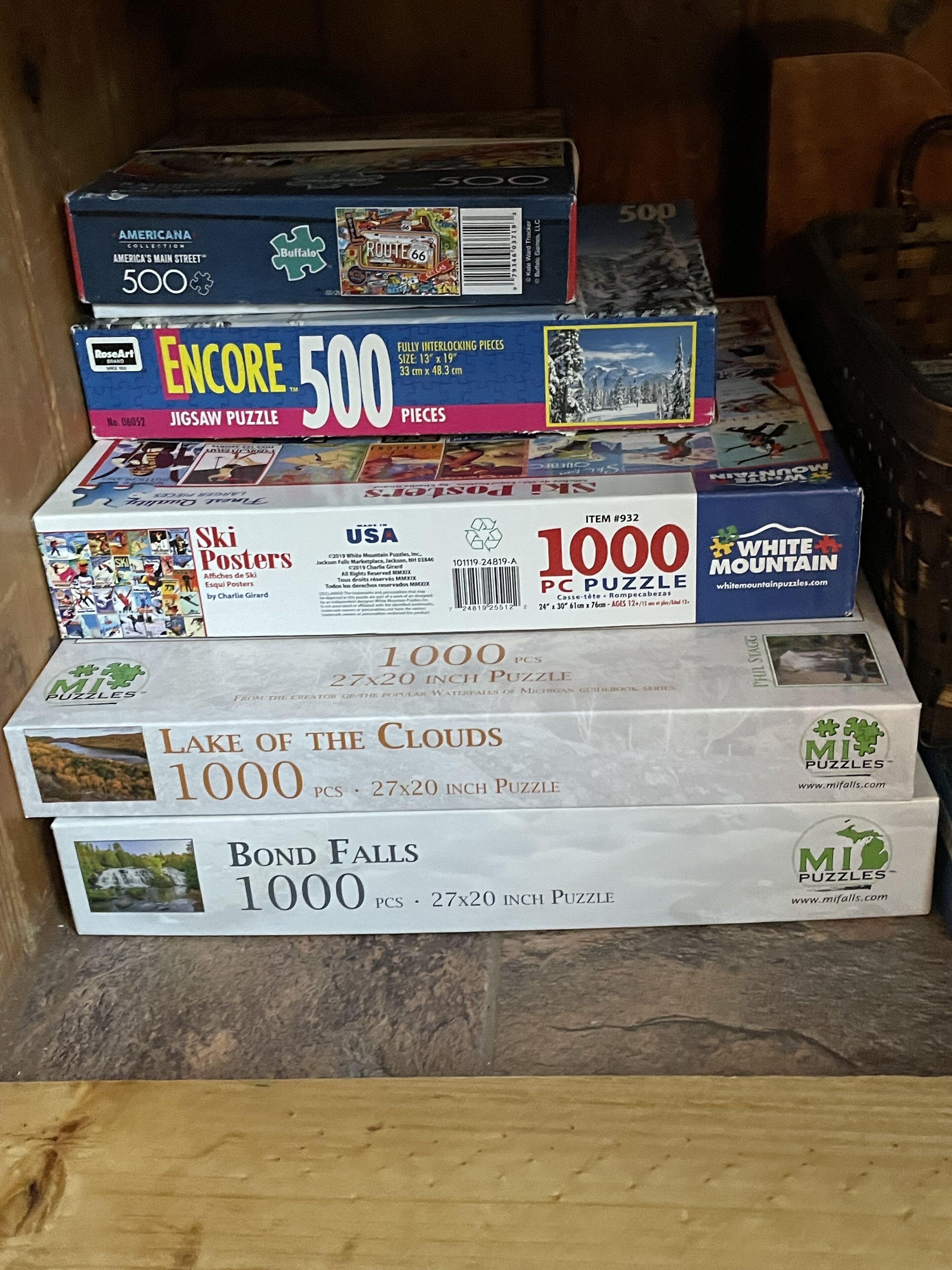 Une pile de jeux de puzzle de différentes difficultés et sur des thèmes variés, comprenant un puzzle de 500 pièces et un puzzle de 1000 pièces.