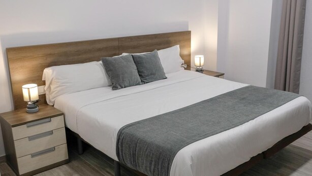 Ein ordentlich bezogenes Bett mit weißen Leinen und grauen Kissen, an beiden Seiten mit Nachttischen und Lampen, in einem modernen Zimmer mit Holz-Kopfstück.