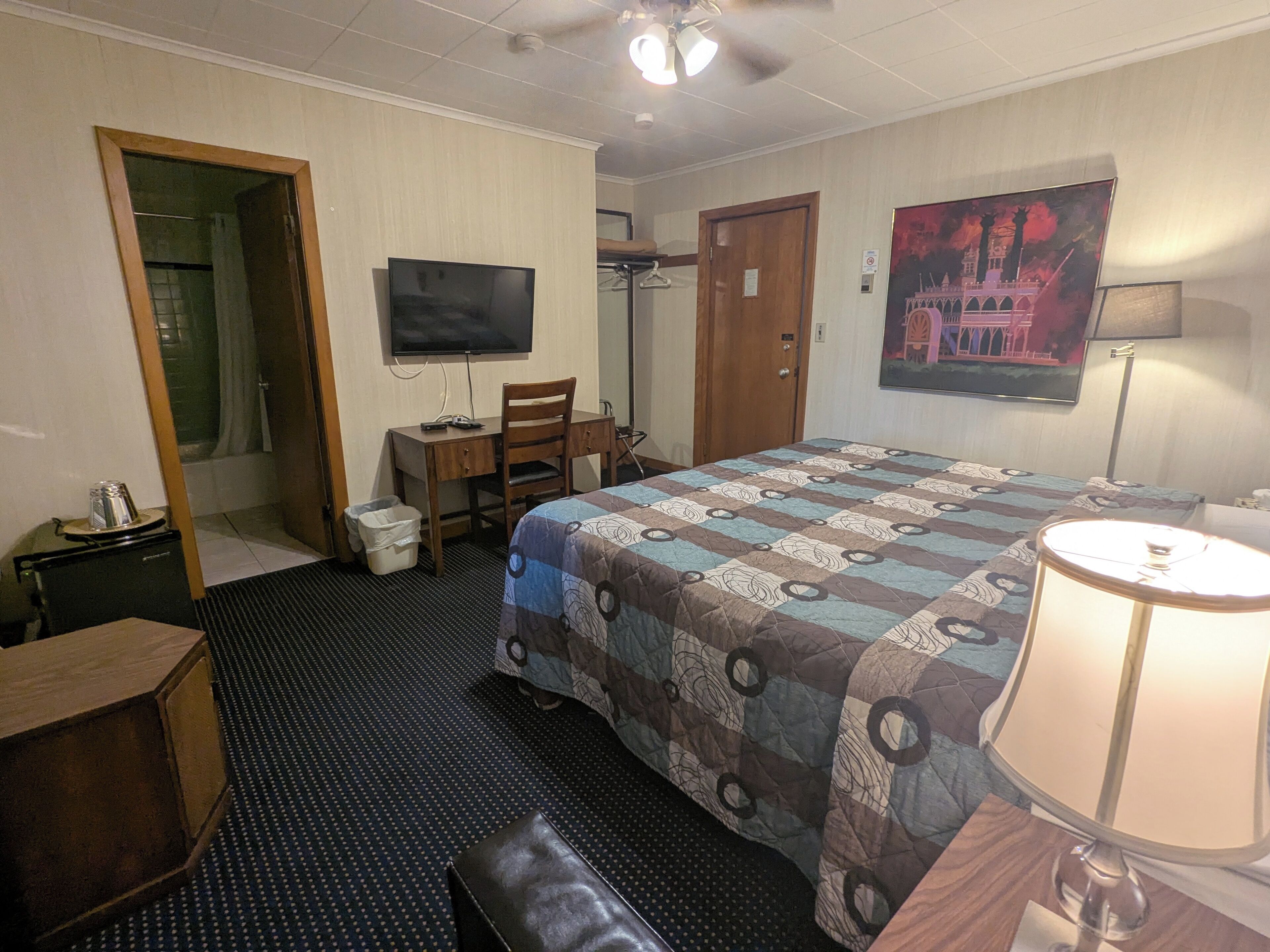 A hotel room with a bed, a desk, a chair, a television, a door, and a painting on the wall.