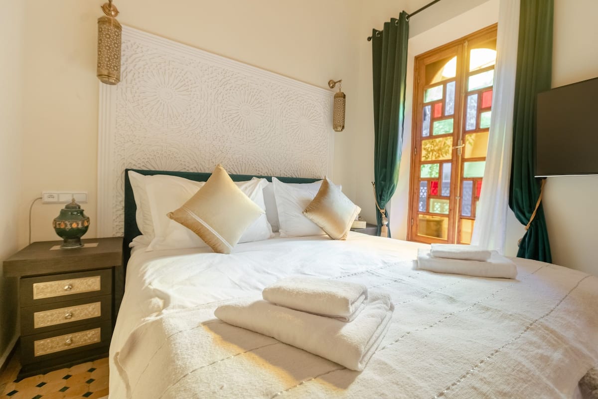 A neatly made bed with pillows and folded towels, a nightstand with a lamp, and a window with curtains.