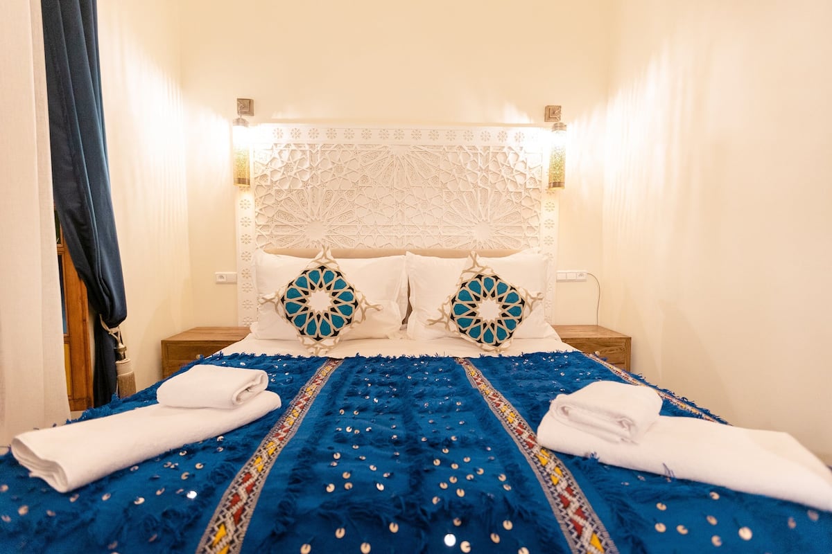 A neatly made bed with a blue bedspread and white pillows, a wooden nightstand, and a decorative headboard.