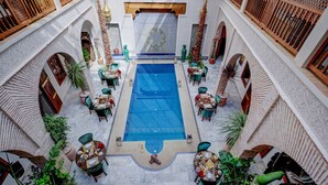 An indoor swimming pool with multiple seating areas and tables, surrounded by arches and potted plants. - Riad Janate (Marrakech)