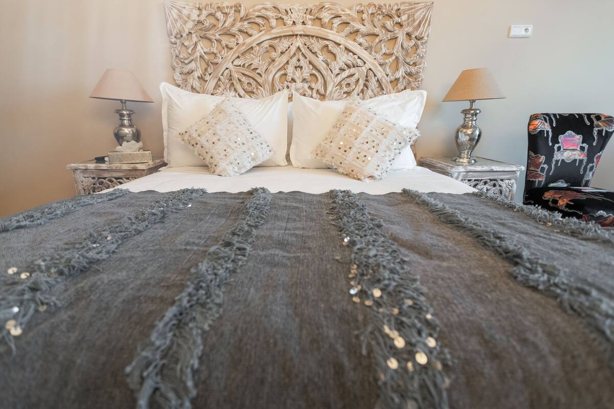 A bed with a decorative headboard, two bedside tables with lamps, and a chair with a patterned seat.