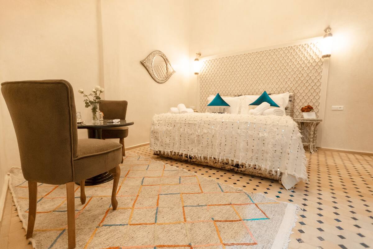 A neatly arranged bedroom with a bed, a chair, a small table, and a decorative mirror.