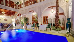 A pool area with a blue floor, surrounded by tables and chairs, and a wooden balcony with intricate railings. - Riad Janate (Marrakech)