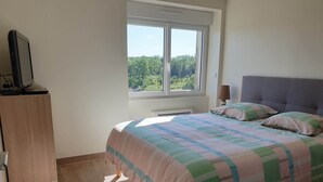 A bedroom with a bed, a desk, and a window with a view of greenery.