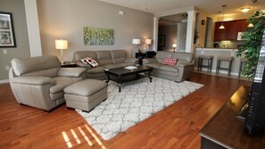 A modern living room with a sectional sofa, a coffee table, and a rug. There is a kitchen area visible in the background.