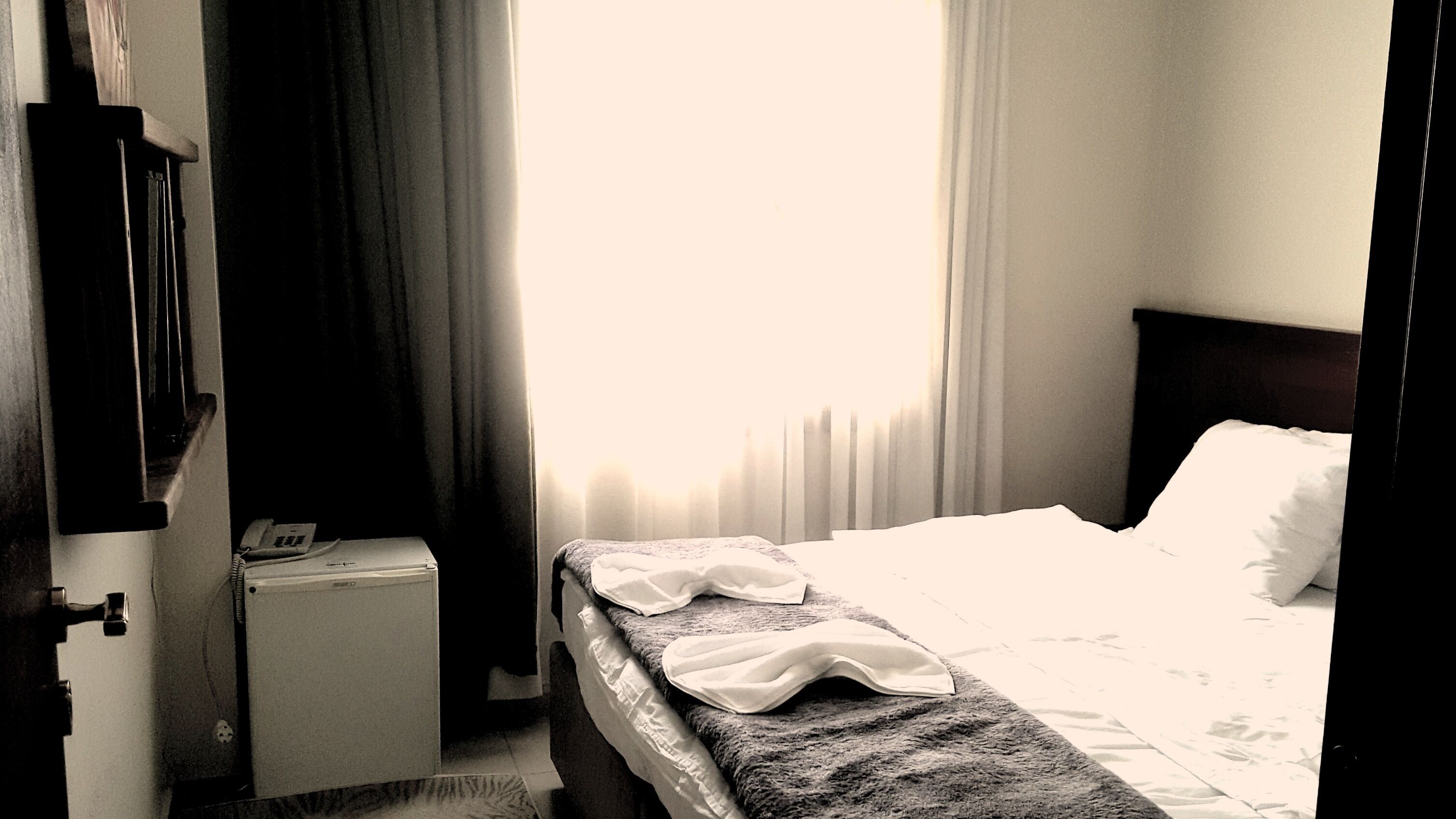 Habitación de hotel con una cama individual, una mesita de noche pequeña con lámpara, una ventana con cortinas y una puerta.