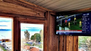 Une pièce d’angle avec des lambris en bois, une grande fenêtre donnant sur un quartier résidentiel et un téléviseur fixé au mur diffusant un match de sport.