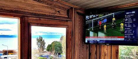 Ein Eckzimmer mit Holzvertäfelung, einem großen Fenster mit Blick auf eine Wohngegend und einem Wandfernseher, der ein Sportspiel zeigt.
