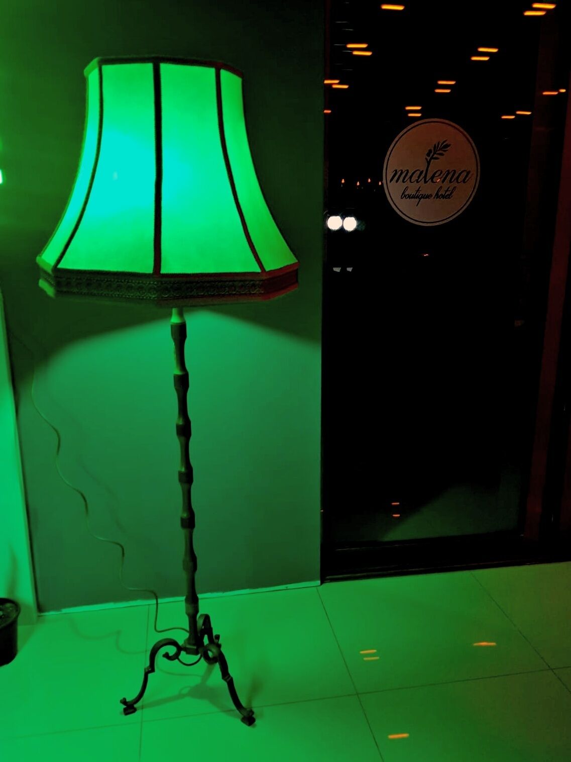Una lámpara de pie verde con una base decorativa, ubicada frente a un cartel que dice 'Malena Boutique Hotel'.