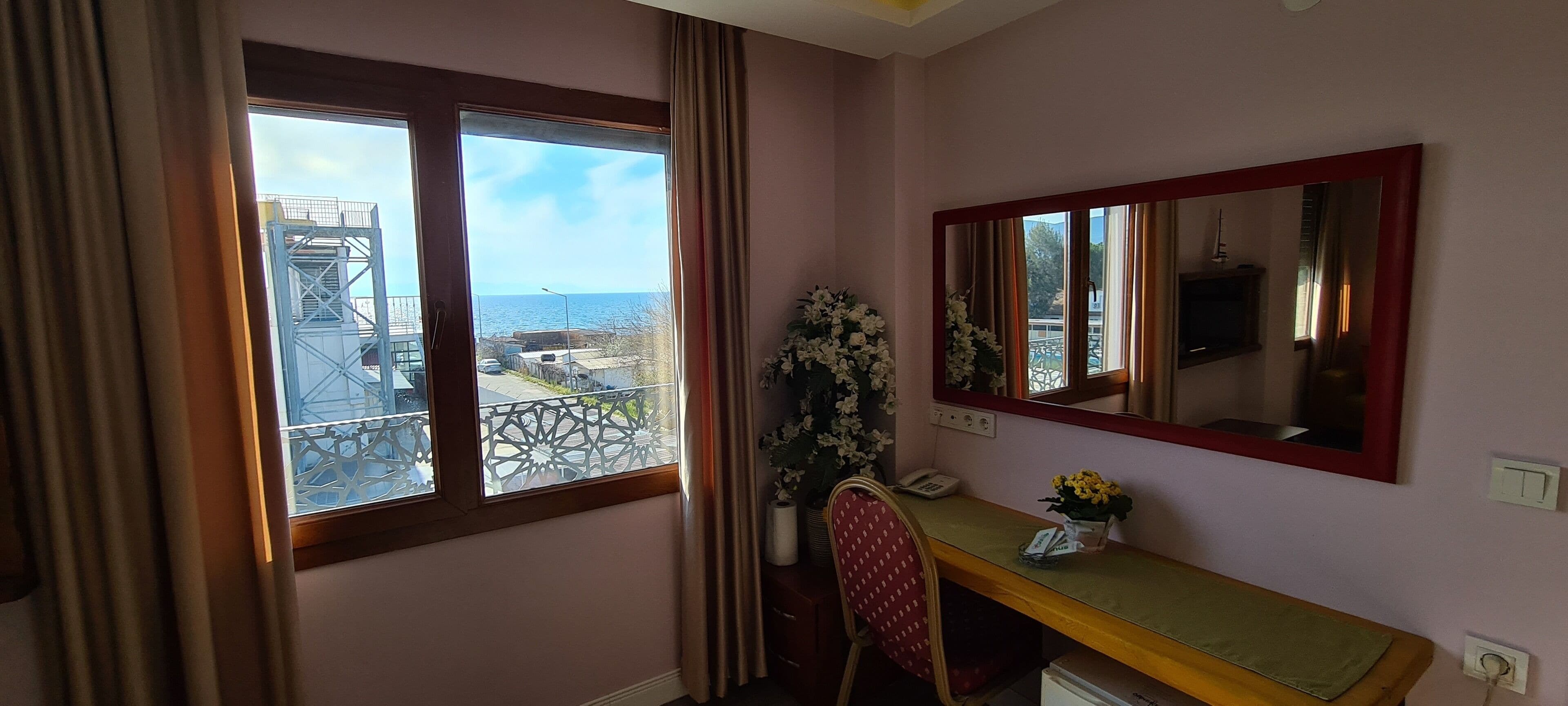 A room with a desk, chair, and a large mirror. There is a window with a view of a balcony and the sea.