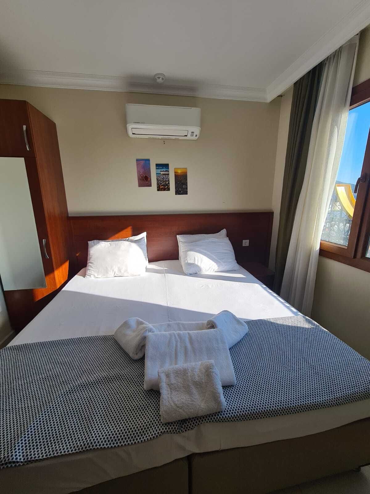 A hotel room with a bed, an air conditioning unit, a window with curtains, and a wall with three pictures.