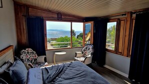 Une chambre avec un grand lit, une chaise, une petite table et une vue sur la mer grâce à de grandes fenêtres.