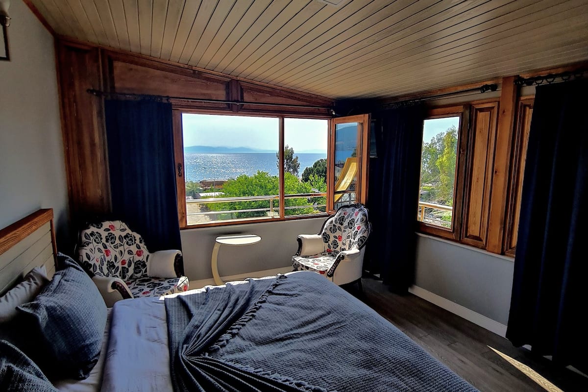 A bedroom with a large bed, a chair, a small table, and a view of the sea through large windows.
