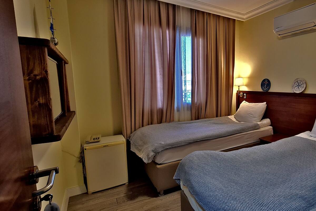 A hotel room with two beds, a wooden headboard, a small bedside table, a nightstand, a wall-mounted air conditioner, and a window with curtains.