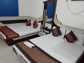 A hotel room with two beds, each with a white mattress, brown headboard, and decorative pillows. There is a small bedside table with a lamp and a power outlet.