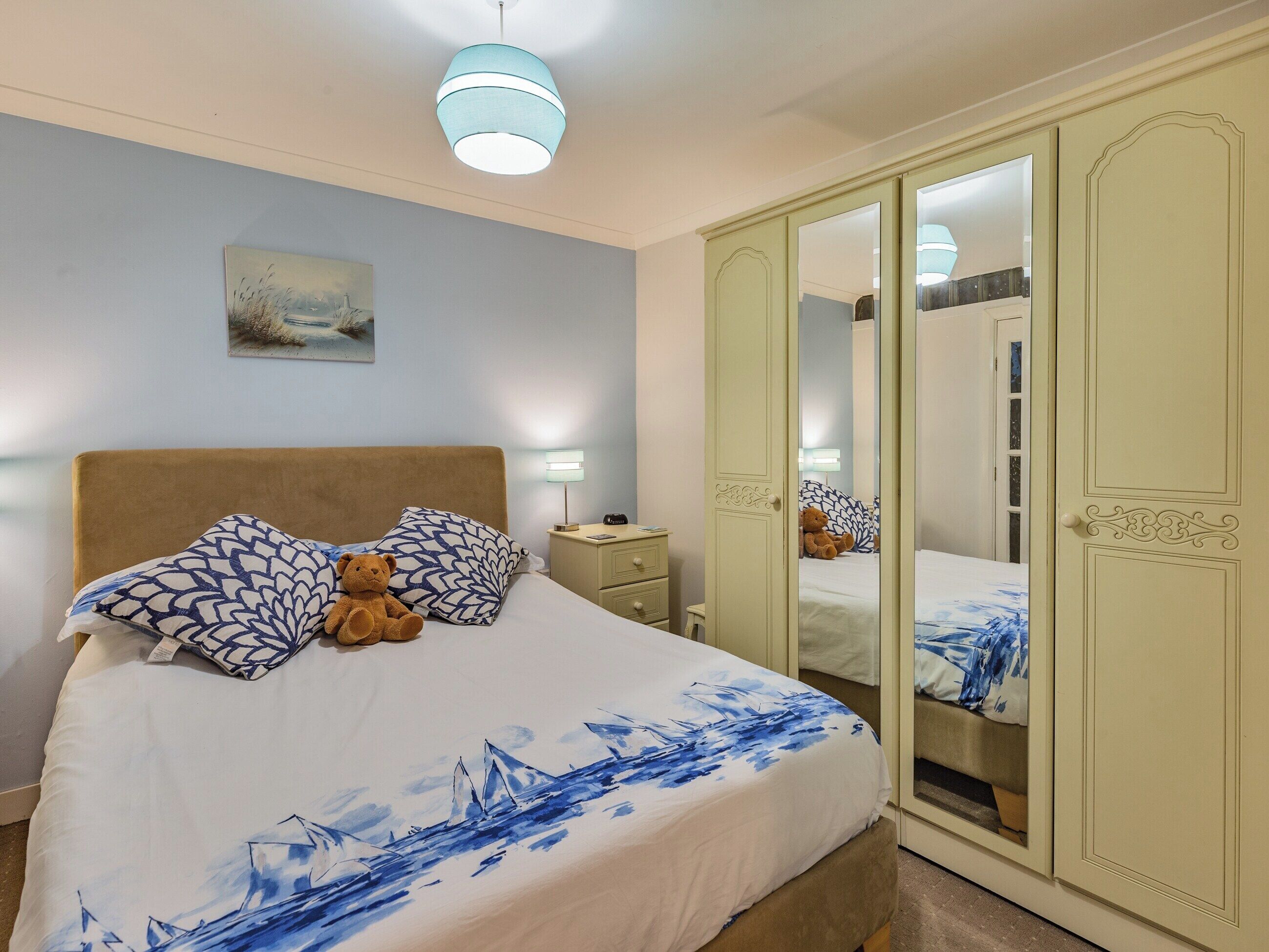 A bedroom with a bed, pillows, a teddy bear, a bedside lamp, a nightstand, and a wardrobe with a mirror.