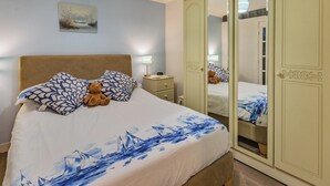 Un dormitorio con cama, almohadas, un oso de peluche, una lámpara de noche, una mesita de noche y un armario con espejo.