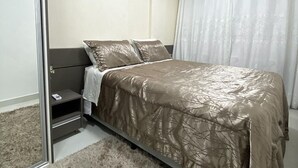 Un dormitorio con cama, mesita de noche, espejo y ventana con cortinas.