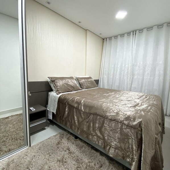 A bedroom with a bed, a nightstand, a mirror, and a window with curtains.