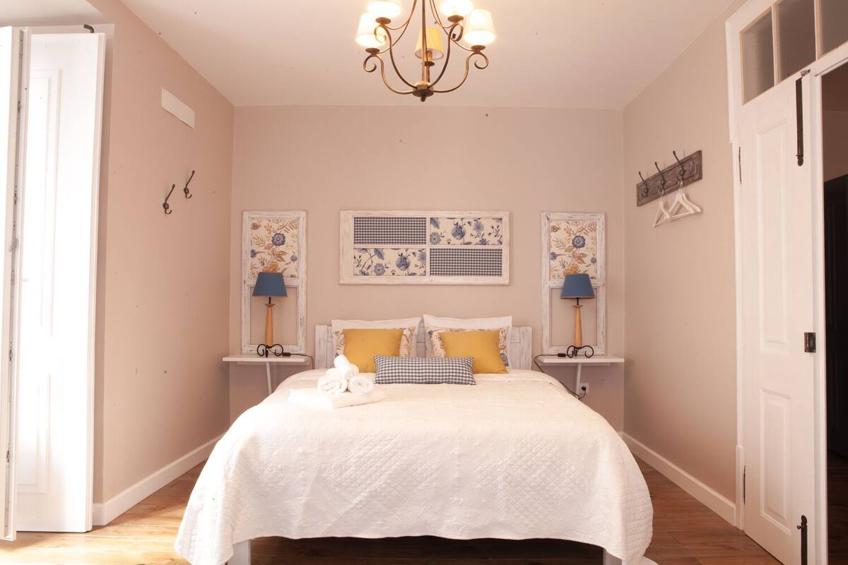 A bedroom with a bed, two bedside tables, a chandelier, and floral-patterned wall art.