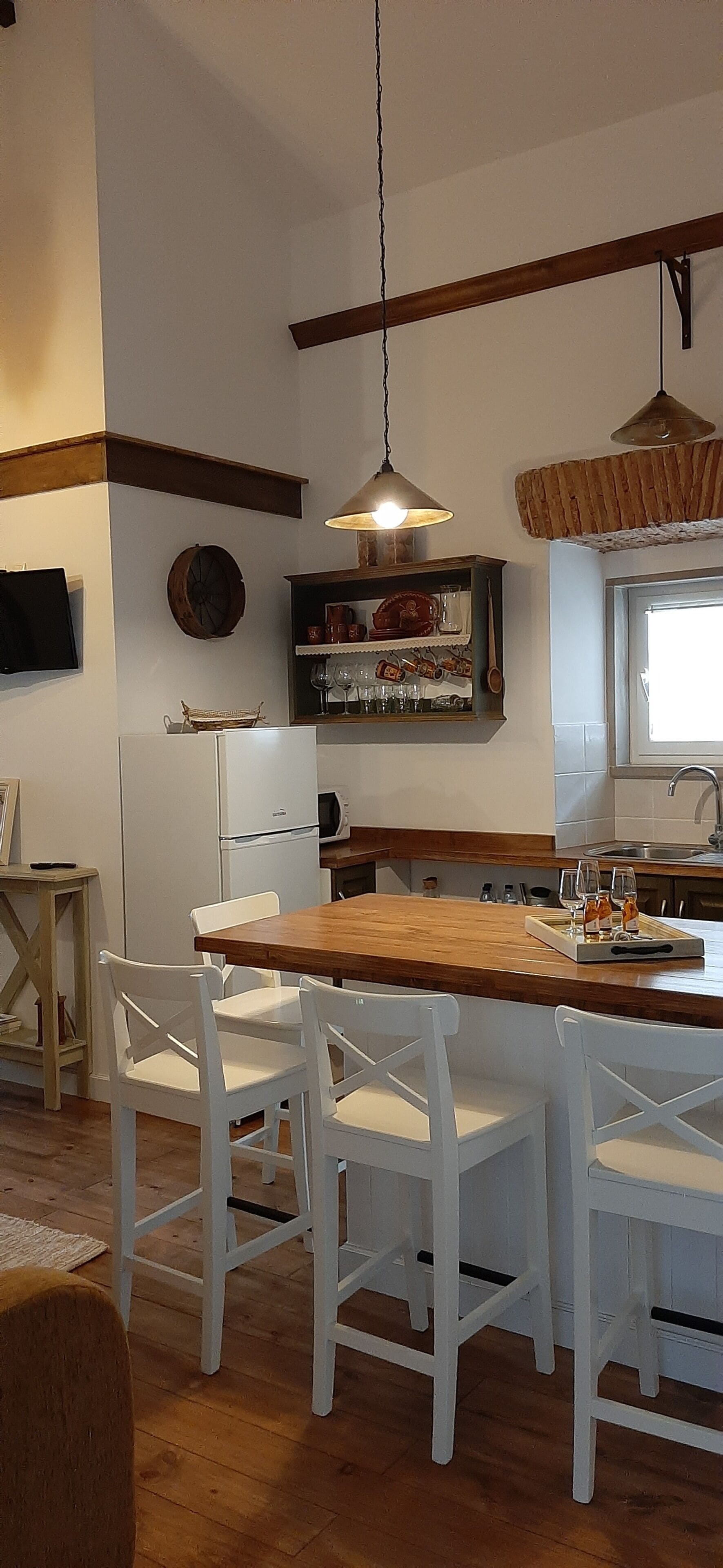 Uma cozinha com mesa de madeira e cadeiras brancas, geladeira, micro-ondas e luminária pendente.
