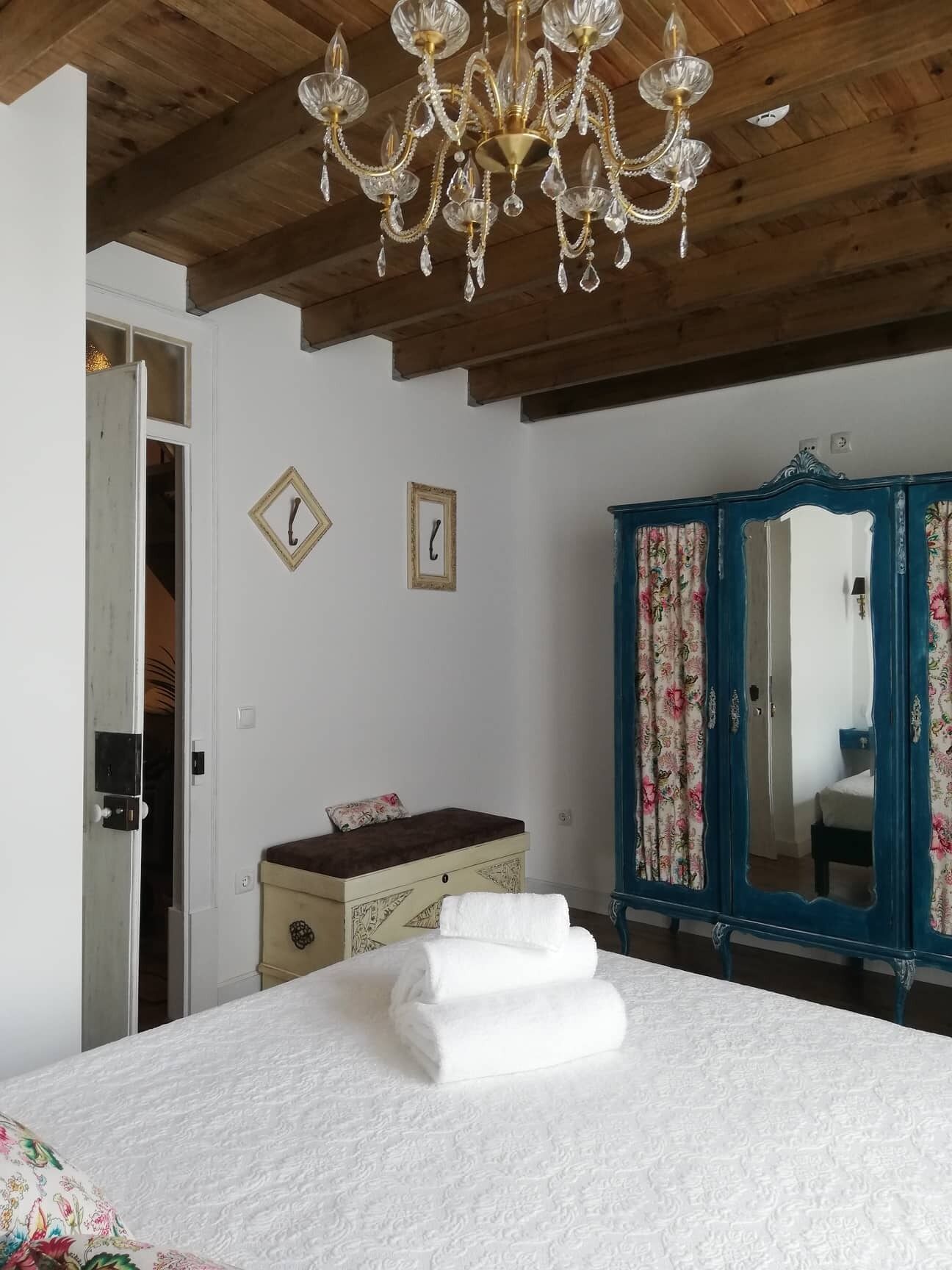 A bedroom with a wooden ceiling, a chandelier, a bed with white bedding, a dresser, and a blue mirror with floral curtains.