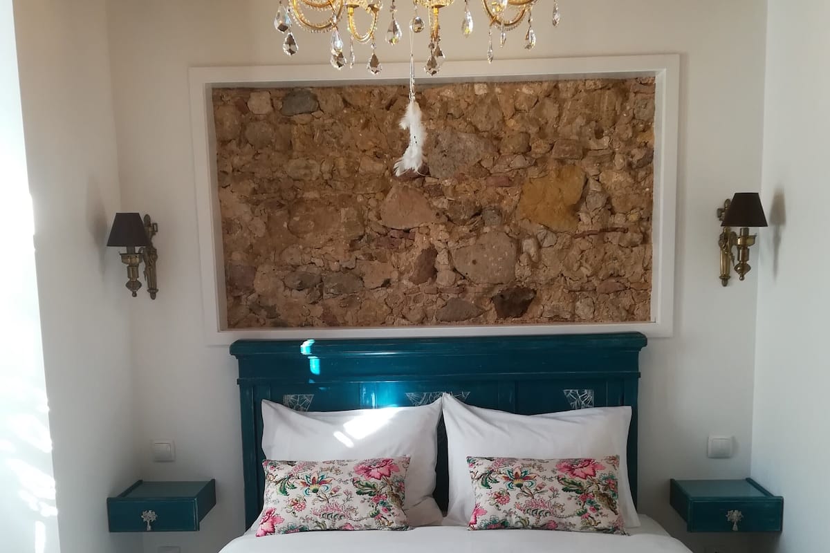 A bedroom with a large bed, floral pillows, a stone wall, a chandelier, and two bedside tables.